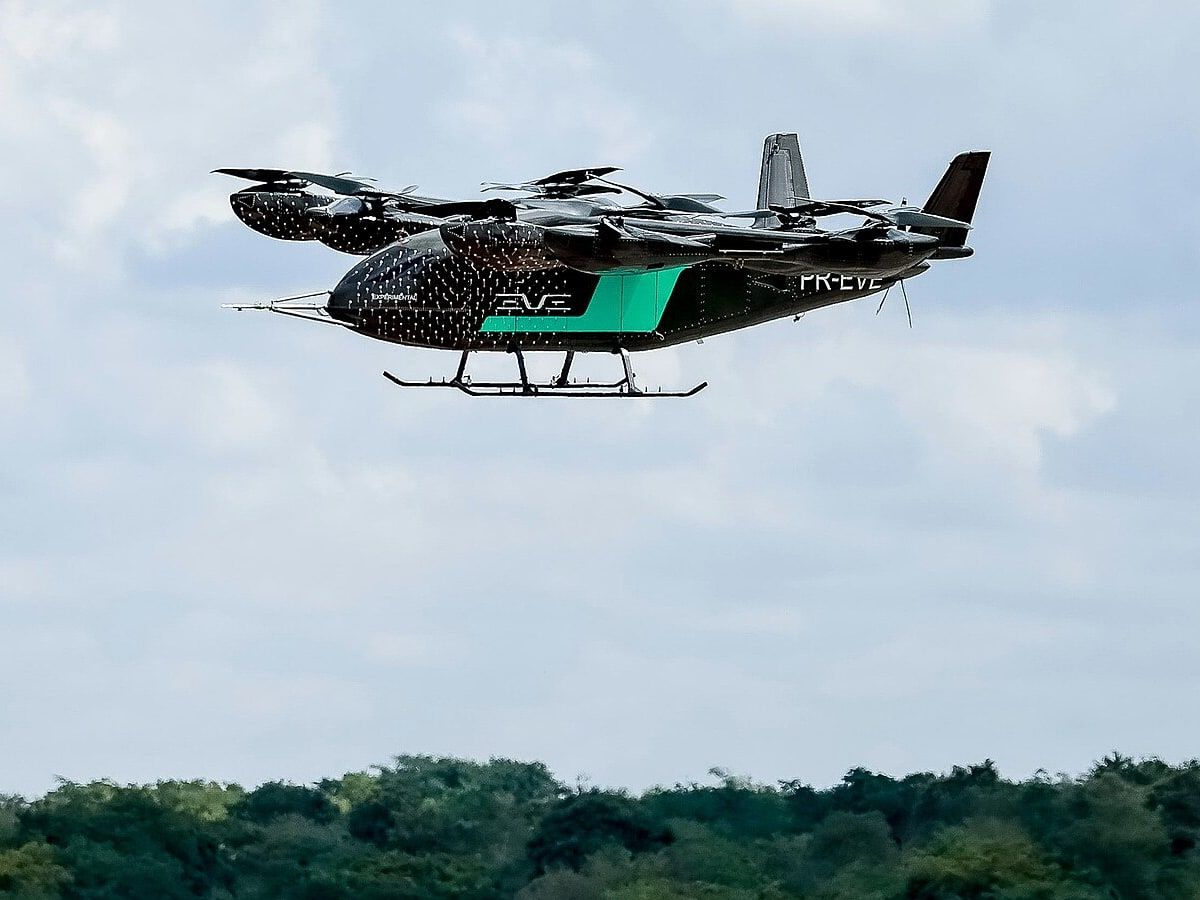 Protótipo do eVTOL da Eve, preto com faixa verde, em voo sobre a pista e a mata