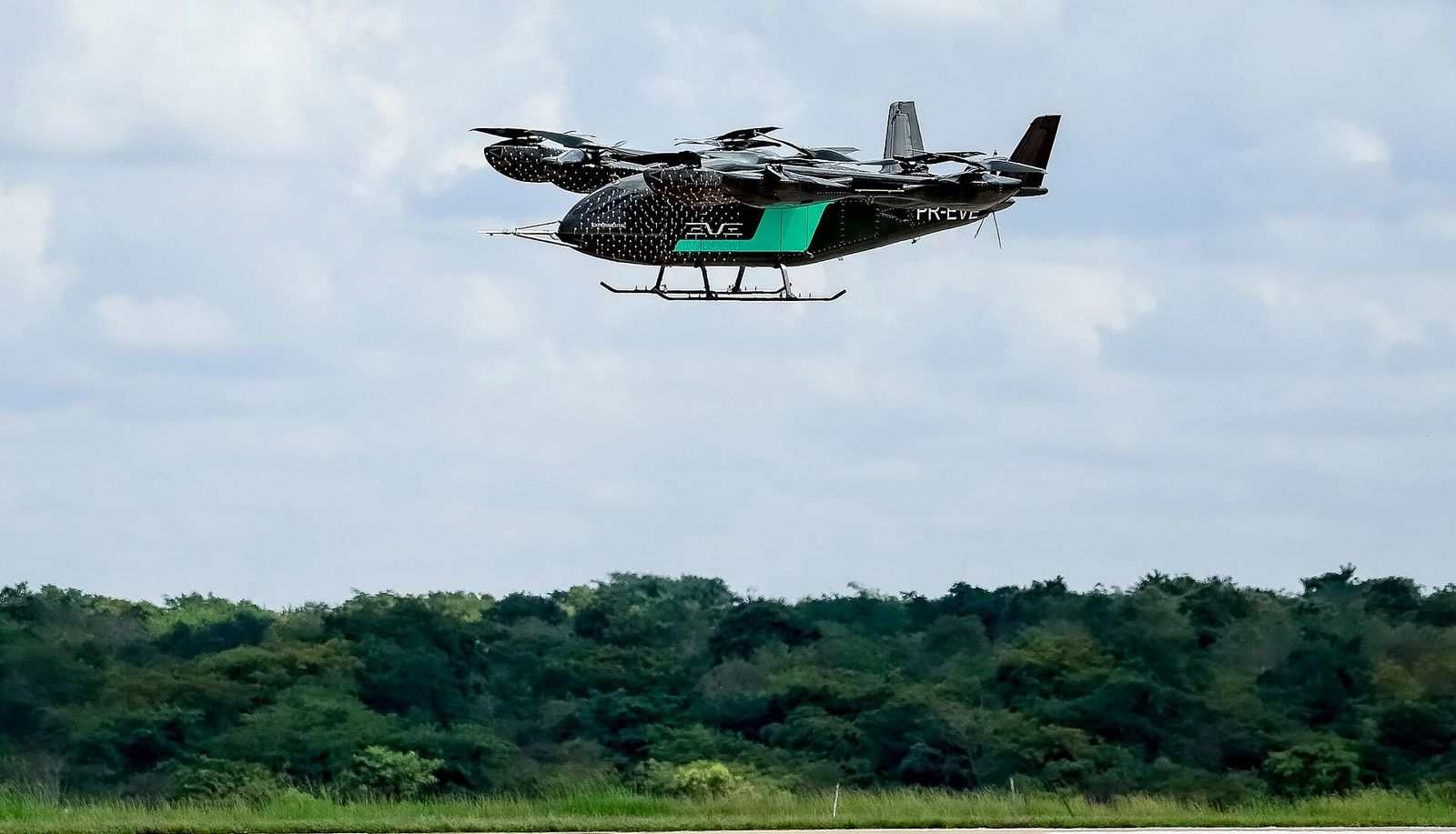 Protótipo do eVTOL da Eve, preto com faixa verde, em voo sobre a pista e a mata