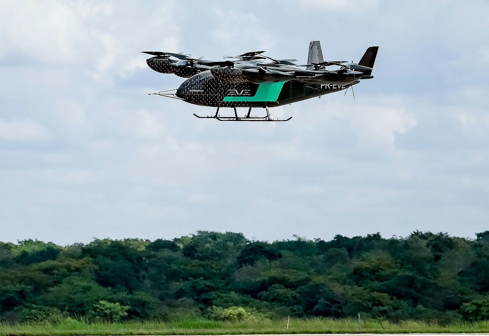 Protótipo do eVTOL da Eve, preto com faixa verde, em voo sobre a pista e a mata