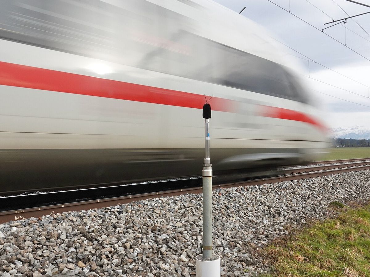 Microfone de medição de ruído no alto de um poste, ao lado dos trilhos, enquanto um trem de alta velocidade passa em movimento