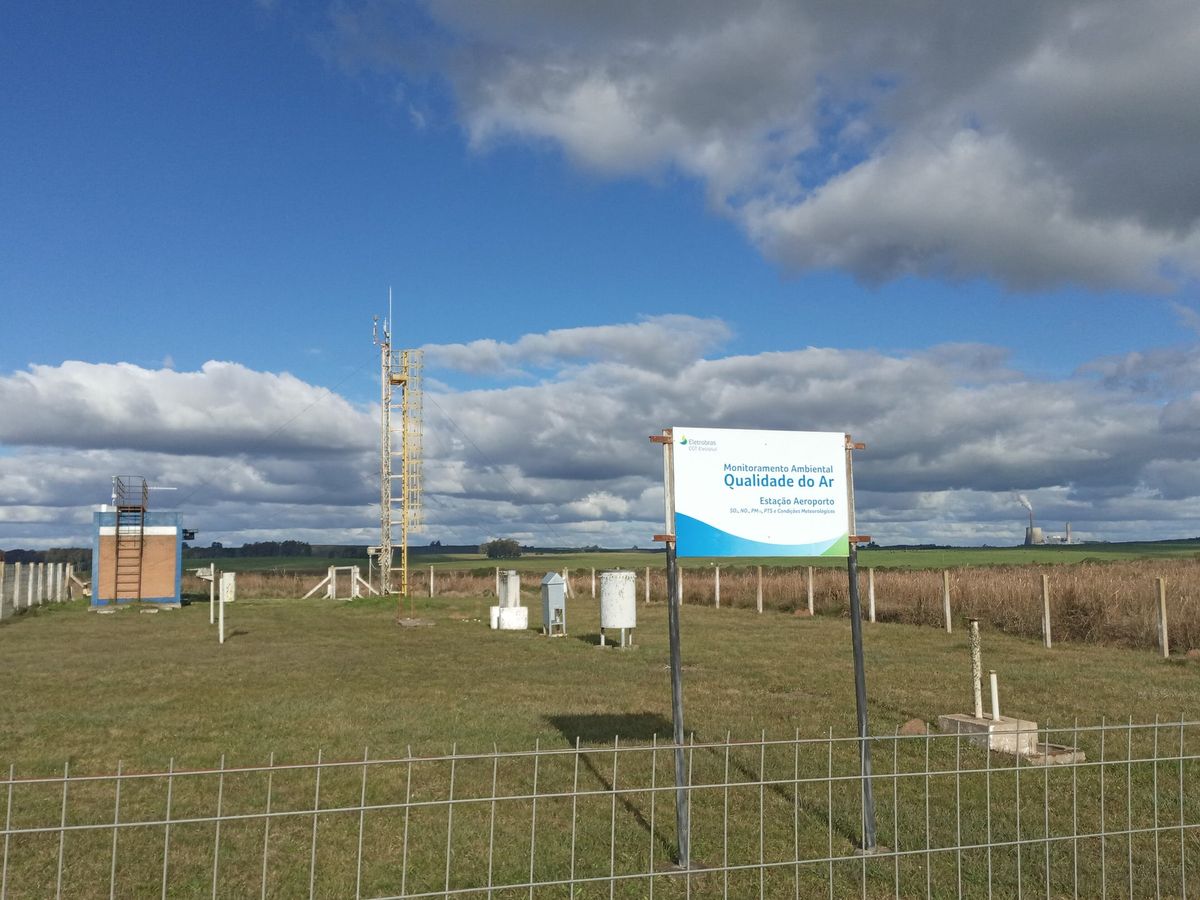 Estação de monitoramento da qualidade do ar em campo aberto, com torre meteorológica, amostradores e a placa da estação, sob céu com nuvens